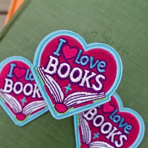 May include: Three heart-shaped embroidered patches featuring a magenta background with a light blue border. Each patch displays an open book graphic in white and magenta with the text I love BOOKS in white and light blue lettering.