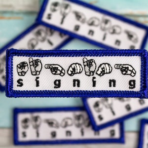 May include: A set of blue and white embroidered patches with the word "signing" spelled out in American Sign Language.