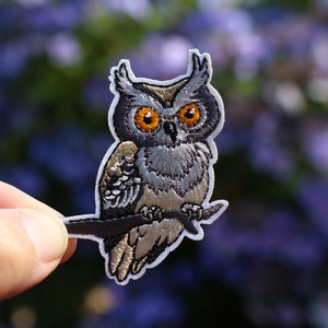 May include: Embroidered iron-on patch of an owl with orange eyes, perched on a branch. The owl is gray and brown with detailed feathers.