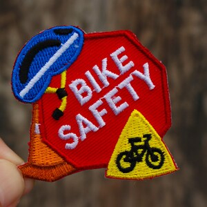 May include: A red and white embroidered patch with the words "BIKE SAFETY" in white. The patch also features a blue helmet, a yellow and orange bicycle, and a yellow triangle with a black bicycle inside.