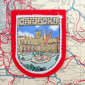 May include: Embroidered patch featuring Chester Cathedral, with gold and blue details, set against a red border. The patch is placed on a vintage map, showcasing a detailed illustration of the cathedral and the text "CHESTER CATHEDRAL".