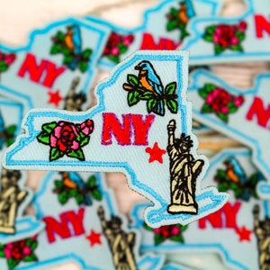 May include: Light blue embroidered patch in the shape of New York State. The patch features the letters "NY" in red, a red star, a rose, a bird, and the Statue of Liberty. The patch has a light blue border.