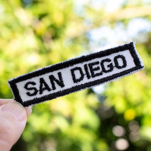 May include: A rectangular patch with the text "SAN DIEGO" embroidered in white on a black and white background. The patch is held against a blurred green background.
