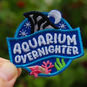 May include: Blue and white embroidered patch with the text "Aquarium Overnighter" and a cartoon shark fin and moon. The patch has a white outline and is surrounded by a blue border with white snowflakes and green and pink sea life.