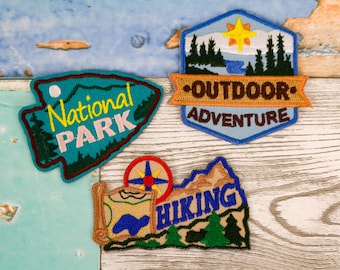May include: Embroidered patches with outdoor themes. Designs include "National Park" with a mountain and moon, "Outdoor Adventure" with trees and a sun, and a "Hiking" patch with a map and compass. Patches are in shades of blue, green, yellow, and brown.
