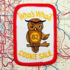 May include: Embroidered patch with a red border and white background. The patch features a winking owl with the text "Who's Who" and "Cookie Sale" in yellow. The owl has the number "82" on its chest. The patch is on a vintage map.
