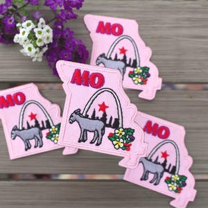 May include: Four light pink embroidered patches shaped like the state of Missouri. Each patch features the red text MO, a grey donkey, a white arch outline, a red star, and a small cluster of colorful flowers in the bottom right corner.