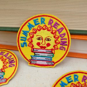 May include: A circular embroidered patch with a yellow background featuring a smiling sun face above a stack of books. The text "SUMMER READING" is stitched in blue and purple around the sun. The patch has a red border and detailed stitching.