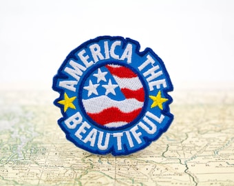 May include: Embroidered patch with the text "AMERICA THE BEAUTIFUL" in white on a blue background. The patch features a circular design with the American flag and two yellow stars. The patch is placed on a vintage map.