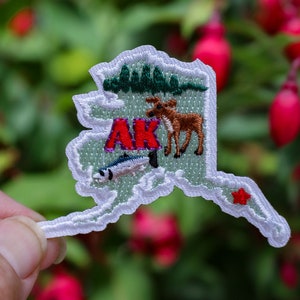May include: White felt patch shaped like the state of Alaska with a red "AK" in the center, a green forest, a brown moose, a white fish, and a red star in the lower right corner.