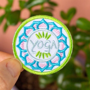 May include: A white embroidered patch with a green outer border and a blue and pink mandala design. The word "yoga" is written in silver script inside the mandala.