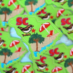 May include: A green felt patch in the shape of South Carolina with the letters "SC" in red, a brown bird, a red and white beach umbrella, a green palm tree, and a red beach chair.