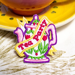 May include: A white embroidered iron-on patch with a pink teapot design and a yellow banner that reads "Tea Party".