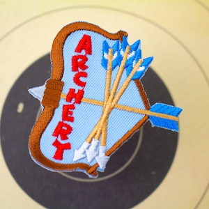 May include: A blue and brown embroidered patch with the word "ARCHERY" in red lettering. The patch depicts a bow and arrows.
