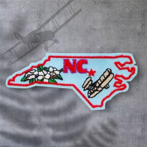 May include: A blue and red embroidered patch shaped like the state of North Carolina. The patch features a white flower, a red star, the letters "NC" in red, and a brown biplane.