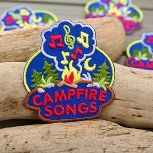 May include: A blue embroidered patch featuring a campfire with red and yellow flames, green pine trees, and a cloud shape containing red, green, and yellow musical notes. The text CAMPFIRE SONGS is embroidered in red letters at the bottom.
