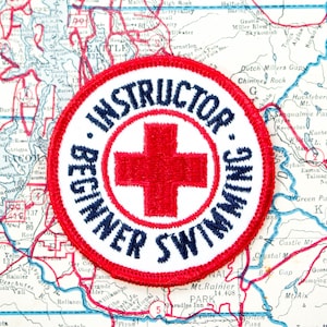May include: A circular embroidered patch featuring a red cross in the center on a white background. The text "INSTRUCTOR" is arched above the cross, and "BEGINNER SWIMMING" is arched below, all in dark blue thread with a red stitched border.