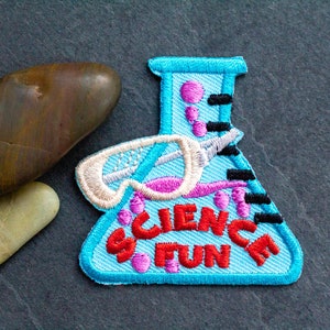 May include: A light blue embroidered patch featuring a science beaker design with the words "SCIENCE FUN" in red. The patch also includes a pair of goggles and black and pink accents. Two stones are next to the patch.