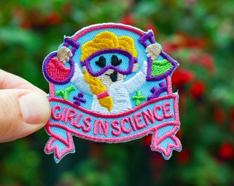 May include: A blue and pink embroidered patch with a cartoon girl wearing goggles and holding beakers. The text "Girls in Science" is on a pink banner.