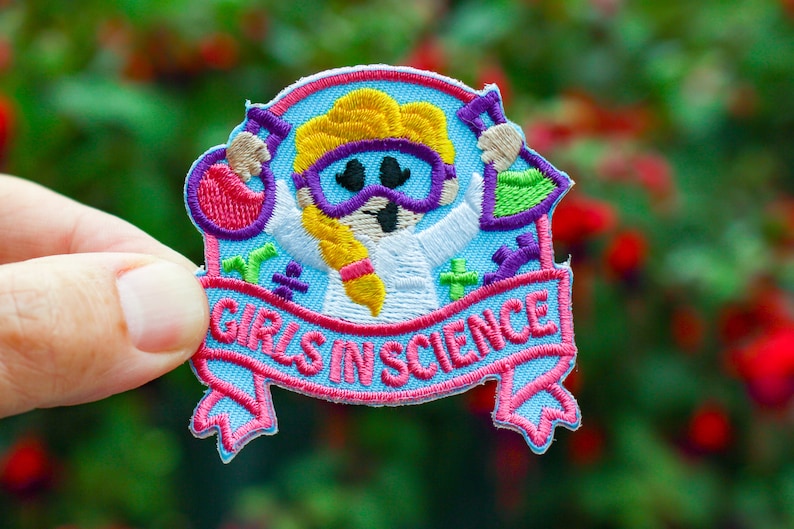 Girls in Science embroidered patch with girls in science in pink thread in a banner and a girl holding lab equipment in safety goggles