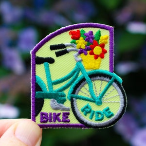 May include: A green and blue bicycle with a basket of flowers on the handlebars. The bicycle has a purple border and the words "Bike Ride" are embroidered on the patch.