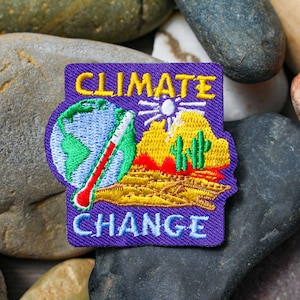 May include: Purple embroidered patch with the words "CLIMATE CHANGE" in yellow lettering. The patch features a globe with a red thermometer, a desert scene with cacti, and a sun.