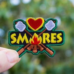May include: A green, yellow, and red embroidered patch with the word "SMORES" and a heart, campfire, and crossed sticks with marshmallows.