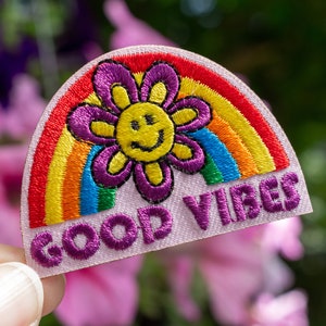 May include: A pink fabric patch with a rainbow and a purple flower with a yellow smiley face. The text "GOOD VIBES" is embroidered in purple.
