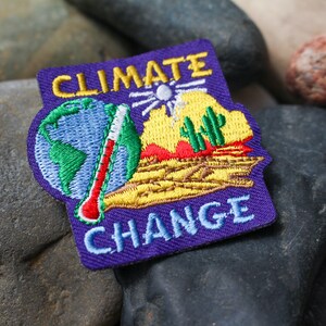 May include: Purple embroidered patch with the text "CLIMATE CHANGE" in yellow and blue lettering. The patch features a globe, a thermometer, a sun, and a desert landscape with cacti.