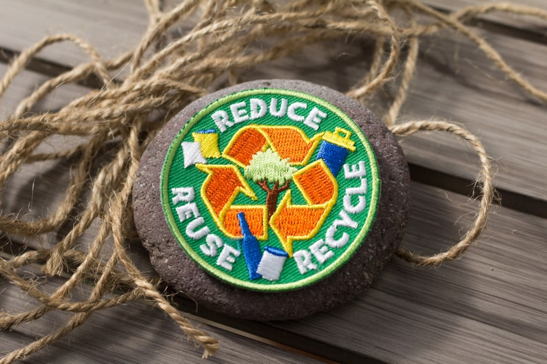 May include: A green, orange, and white embroidered patch with the words "Reduce, Reuse, Recycle" and a stylized recycling symbol with a tree in the center. The patch is attached to a round, dark gray stone.