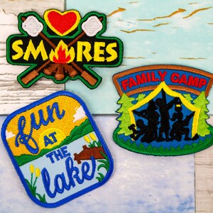 May include: Three embroidered patches with a camping theme. The first patch is yellow and blue with the text "fun at the lake". The second patch is green and blue with the text "family camp" and a silhouette of people camping. The third patch is black, red, and white with the text "SMORES" and a heart, marshmallows, and crossed sticks.