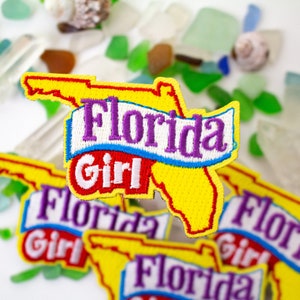 May include: Embroidered patches featuring the outline of Florida in yellow, with the words "Florida Girl" in purple and white. The patches are surrounded by sea glass and seashells, creating a beachy theme.