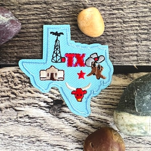 May include: Blue felt outline of the state of Texas with various embroidered details including a cowboy hat, boots, a building, a star, and the word "TX".