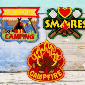 May include: Three embroidered patches with a camping theme. The first patch is blue and green and says "Backyard Camping" with a tent and camping chair. The second patch is green and black and says "S'mores" with a campfire, marshmallows, and sticks. The third patch is red and orange and says "Campfire" with a campfire.