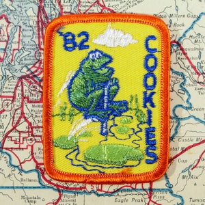 May include: Embroidered patch with a yellow background and an orange border. The patch features a green frog sitting on a log with the text "82 COOKIES" in blue. A white cloud and water are also depicted.