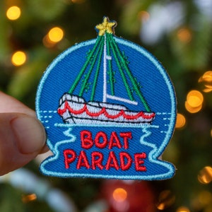 May include: A blue embroidered patch with a white sailboat decorated with red and white trim, a green Christmas tree, and the text "BOAT PARADE".