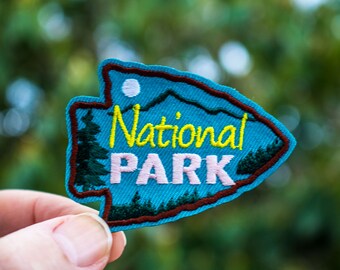 May include: A blue and brown embroidered patch with the text "National Park" in yellow and pink lettering. The patch features a mountain range and pine trees.