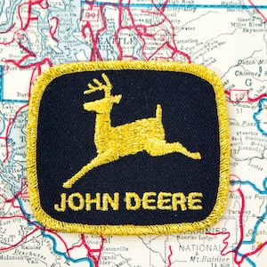 May include: A square embroidered patch with rounded corners featuring a black background and a gold border. The patch displays a gold leaping deer silhouette above the yellow text JOHN DEERE, centered on a detailed map of Washington state.