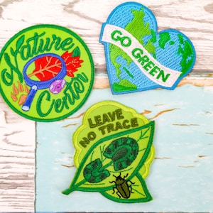 May include: Three green embroidered patches with nature-themed designs. The first patch is round and says "Nature Center" with a magnifying glass and leaves. The second patch is a heart shape with a blue and green globe and says "Go Green". The third patch is a green leaf with a beetle and says "Leave No Trace".