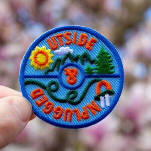 May include: A blue embroidered patch with the text "Outside Unplugged" in orange. The patch features a sun, mountains, a tree, and a tent.