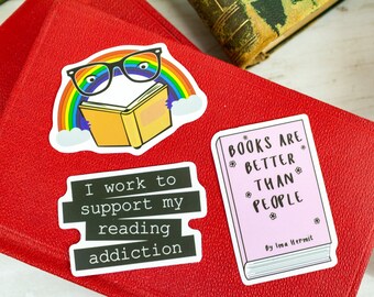 May include: Three book-themed stickers on a red surface. One sticker features a cartoon book wearing glasses and a rainbow. Another sticker says "I work to support my reading addiction". The third sticker is pink and says "Books are better than people" with the text "By Ima Hermit" at the bottom.