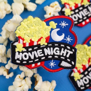 May include: Two embroidered iron-on patches with a movie night theme. One patch features a blue background with a crescent moon and stars, and the other patch features a red, white, and blue striped background with popcorn and the text "MOVIE NIGHT".