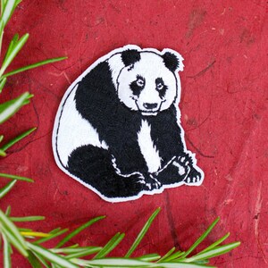 May include: Embroidered panda bear patch with black and white details, sitting on a red textured surface. Green rosemary sprigs are in the foreground. The patch is approximately 3 inches wide.