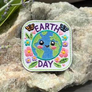 May include: Embroidered patch with a white border featuring a smiling Earth surrounded by colorful flowers and butterflies. The words "EARTH DAY" are embroidered in purple and green. The patch is square-shaped.