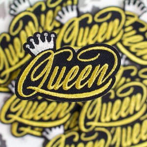 May include: A close-up of several black fabric patches with the word "Queen" embroidered in gold thread. The patches have a silver crown on top.