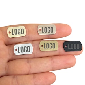 Könnte beinhalten: Vier Metallanhänger mit dem eingravierten Wort "LOGO". Die Anhänger sind in Silber, Gold, Messing und Schwarz.