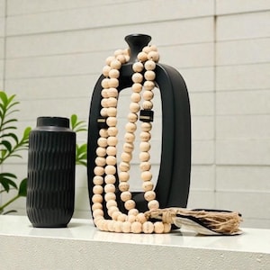 May include: A black, modern vase with a textured surface and a tall, open-center vase with a beaded garland. The garland is made of light-colored wooden beads. A decorative tassel rests on the surface.