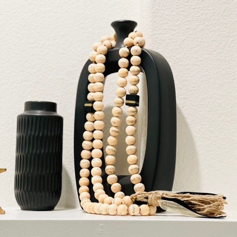 May include: A black ceramic vase with vertical ridges sits next to a tall, black, open-shaped vase. A strand of light-colored wooden beads with etched text hangs from the black vase. A bundle of dried plant material with a decorative tag rests on the shelf.