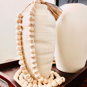 May include: A decorative wooden bead garland with black lettering, draped near a white ceramic vase with a textured surface. A second white vase is in the background. The garland is made of light-colored wooden beads.