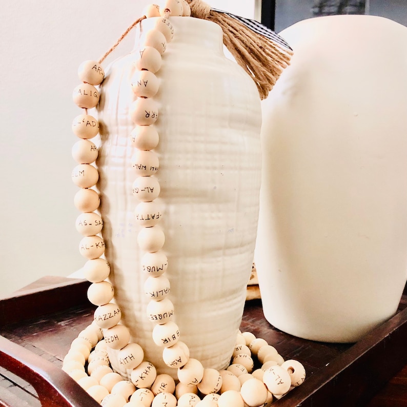 May include: A decorative wooden bead garland with black lettering, draped near a white ceramic vase with a textured surface. A second white vase is in the background. The garland is made of light-colored wooden beads.